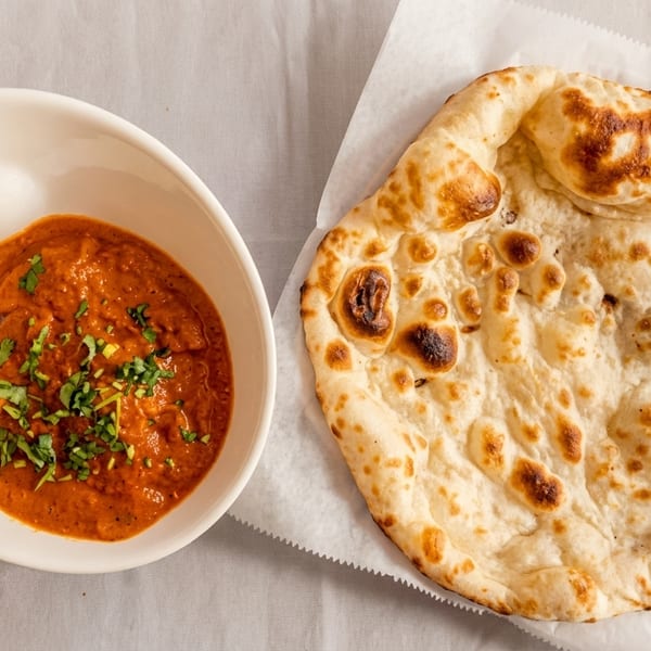 A bowl of orange curry with green herbs next to a piece of naan bread.