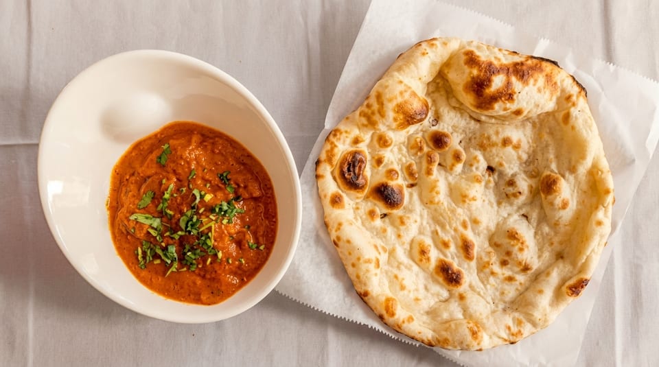 A bowl of orange curry with green herbs next to a piece of naan bread.
