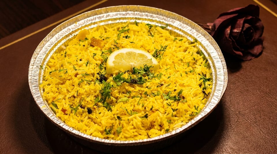 Yellow rice with herbs and a lemon slice in a foil container, next to a dark red rose.